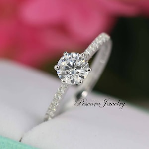 Puede incluir: Primer plano de un anillo de compromiso de diamantes. El anillo presenta un diamante grande, redondo y de talla brillante engastado en una montura de seis puntas. La banda es de metal blanco y está incrustada con diamantes más pequeños.