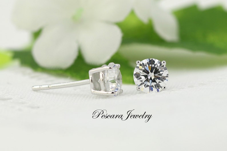 May include: A pair of silver stud earrings with clear round cubic zirconia stones. The earrings are set against a white background with a green leaf and white flower.