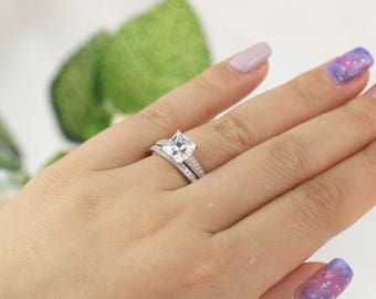 Puede incluir: Un anillo de plata con una gran piedra preciosa transparente de forma cuadrada, rodeada de piedras preciosas transparentes más pequeñas. El anillo se lleva en el dedo anular de la mano izquierda de una persona.