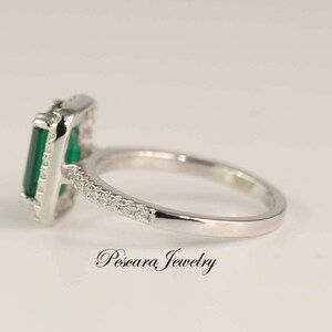 May include: A silver ring with a large emerald green gemstone set in a square frame. The ring is accented with smaller clear gemstones around the frame and band.
