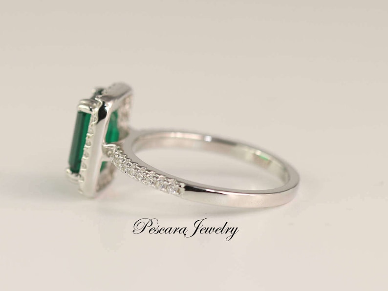 May include: A silver ring with a large emerald green gemstone set in a square frame. The ring is accented with smaller clear gemstones around the frame and band.