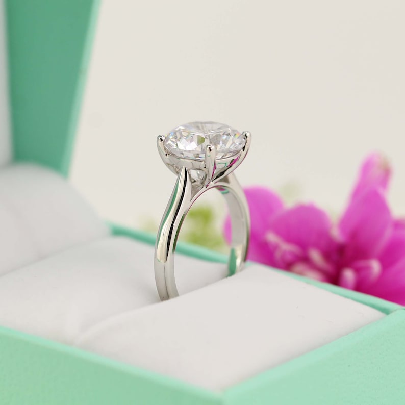 May include: A silver engagement ring with a large, clear, round-cut diamond set in a four-prong setting. The ring is in a light green ring box.