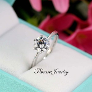 May include: A silver ring with a large, clear, round gemstone in a ring box. The box is lined with white velvet and has a turquoise blue exterior. The text "Pescara Jewelry" is printed on the box.