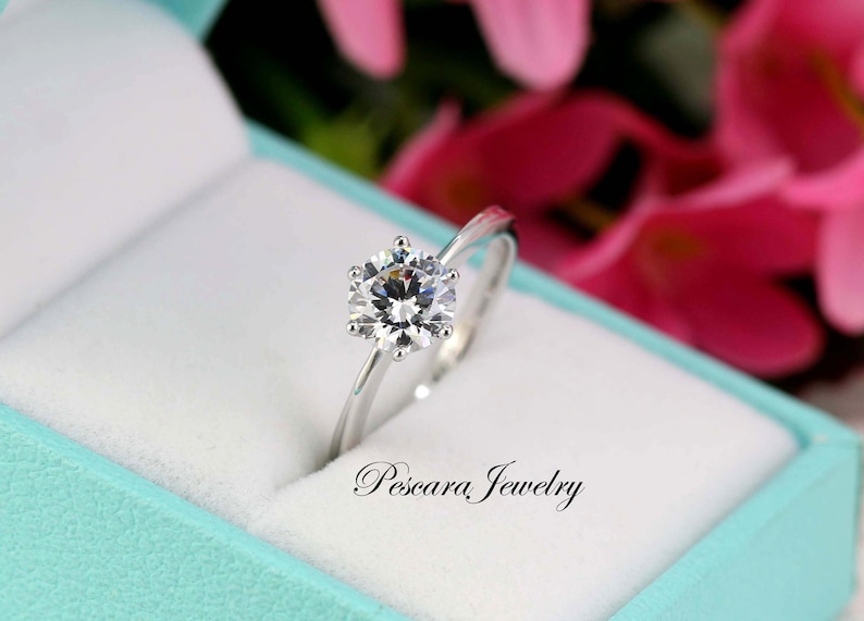 May include: A silver ring with a large, clear, round gemstone in a ring box. The box is lined with white velvet and has a turquoise blue exterior. The text "Pescara Jewelry" is printed on the box.