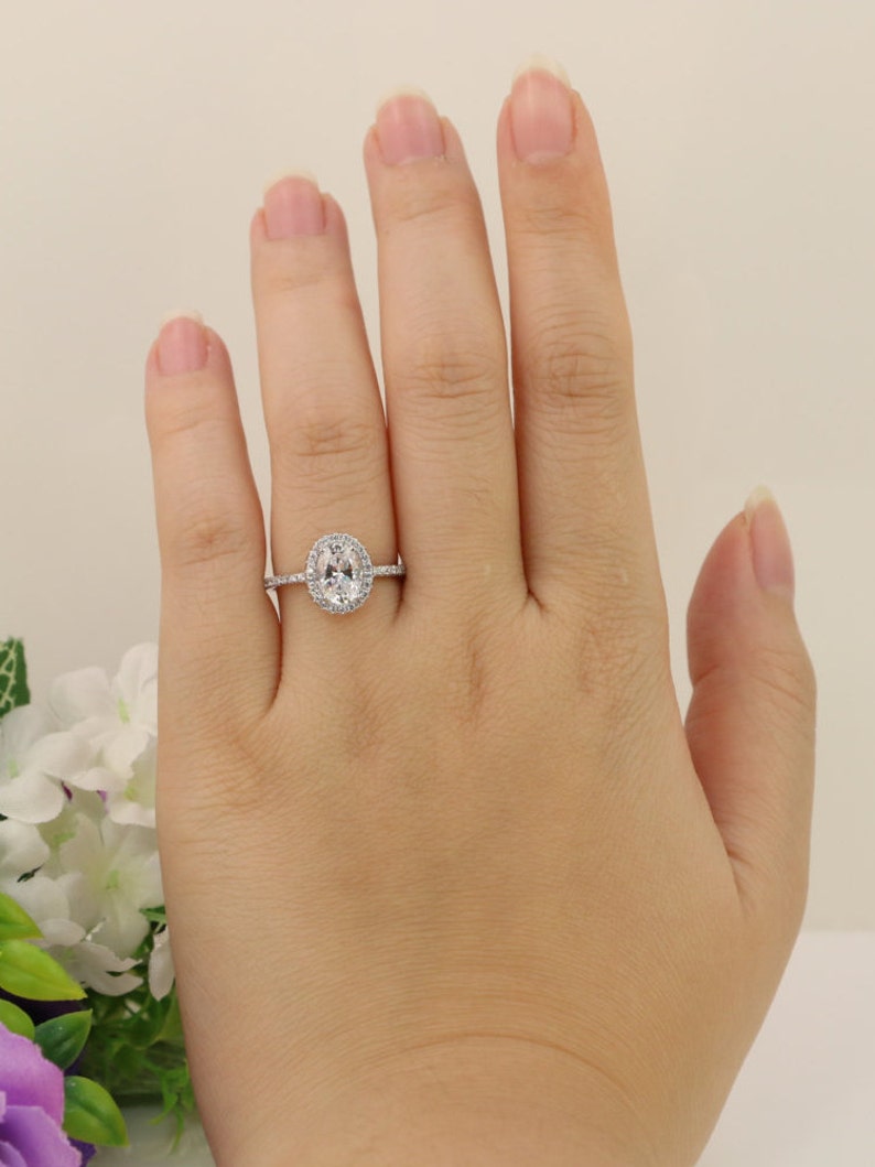 Op de afbeelding: Een zilveren ring met een ovale middelste steen die omringd is door kleinere stenen. De ring zit aan een vinger.