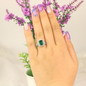 May include: A close-up of a hand wearing a silver ring with a large emerald green gemstone surrounded by smaller diamonds. The ring is on the ring finger of the left hand.