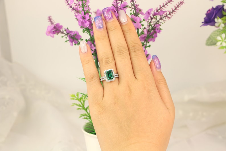 May include: A close-up of a hand wearing a silver ring with a large emerald green gemstone surrounded by smaller diamonds. The ring is on the ring finger of the left hand.