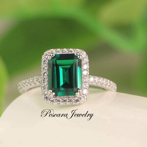 May include: A silver ring with a large emerald green gemstone surrounded by a halo of smaller clear gemstones. The ring is set on a white background.