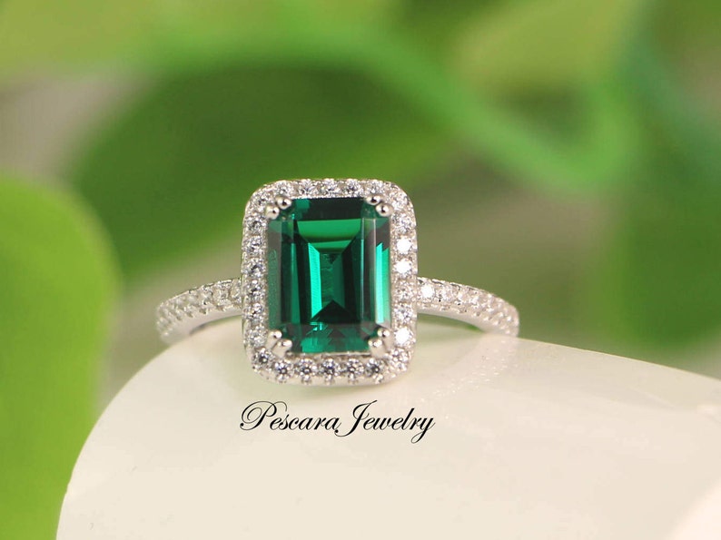 May include: A silver ring with a large emerald green gemstone surrounded by a halo of smaller clear gemstones. The ring is set on a white background.
