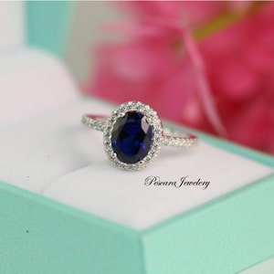 May include: A silver ring with a large oval-shaped blue gemstone surrounded by smaller white gemstones. The ring is in a light blue box with the text "Pescara Jewelry" on the box.