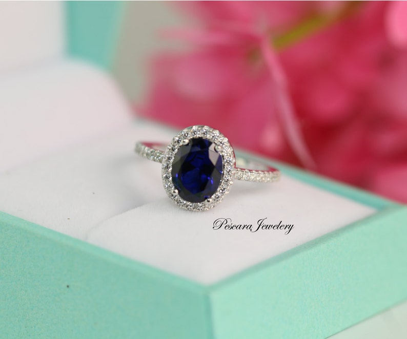 May include: A silver ring with a large oval-shaped blue gemstone surrounded by smaller white gemstones. The ring is in a light blue box with the text "Pescara Jewelry" on the box.