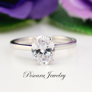 May include: A silver ring with a large oval-shaped clear gemstone. The ring is set on a white background with purple flowers out of focus in the background. The text "Pescara Jewelry" is visible at the bottom of the image.