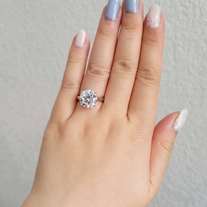 May include: A close-up of a hand wearing a silver ring with a large, round, clear diamond. The ring is on the middle finger of the hand. The hand is wearing light blue and white nail polish.