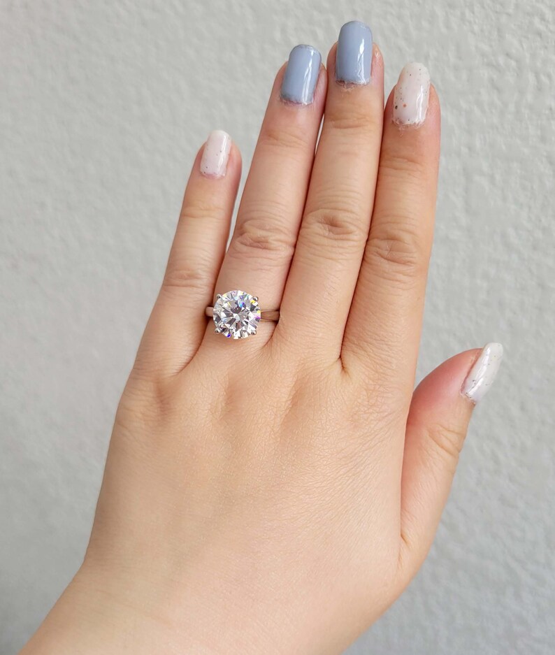 May include: A close-up of a hand wearing a silver ring with a large, round, clear diamond. The ring is on the middle finger of the hand. The hand is wearing light blue and white nail polish.