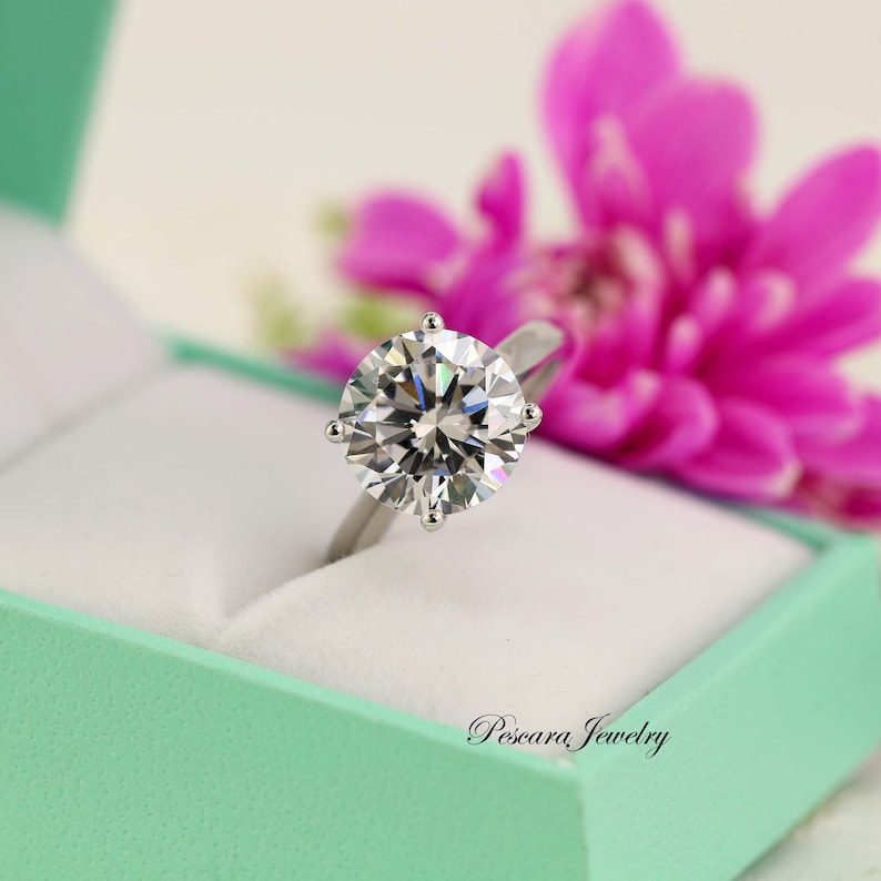 May include: A silver engagement ring with a large, round, clear diamond in a mint green ring box. The ring is set in a six-prong setting. The box has the words "Pescara Jewelry" printed on the front.