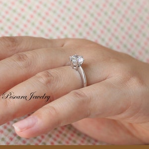 May include: A silver ring with a single, round, clear gemstone set in a prong setting. The ring is on a person's finger.