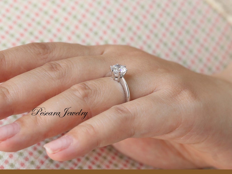 May include: A silver ring with a single, round, clear gemstone set in a prong setting. The ring is on a person's finger.