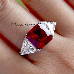 May include: A silver ring with a large, red, square-shaped gemstone surrounded by three smaller, clear, triangular gemstones. The ring is on a finger.