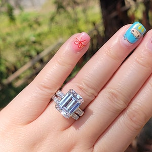 Puede incluir: Un anillo de plata con una gran piedra preciosa transparente de talla esmeralda rodeada de piedras preciosas transparentes más pequeñas. El anillo está en un dedo con esmalte de uñas rosa y blanco.