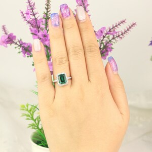 May include: A silver ring with a large emerald green gemstone surrounded by smaller diamonds. The ring is on a hand with painted fingernails.