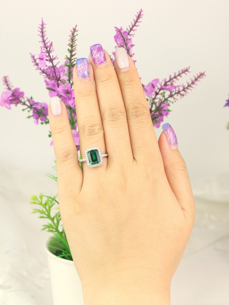May include: A silver ring with a large emerald green gemstone surrounded by smaller diamonds. The ring is on a hand with painted fingernails.