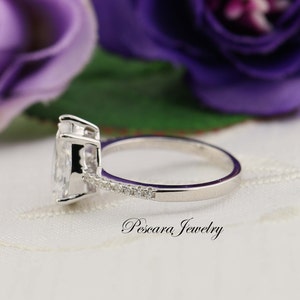 May include: A silver ring with a pear-shaped clear stone and a band of smaller clear stones. The ring is on a white surface with a purple flower in the background.