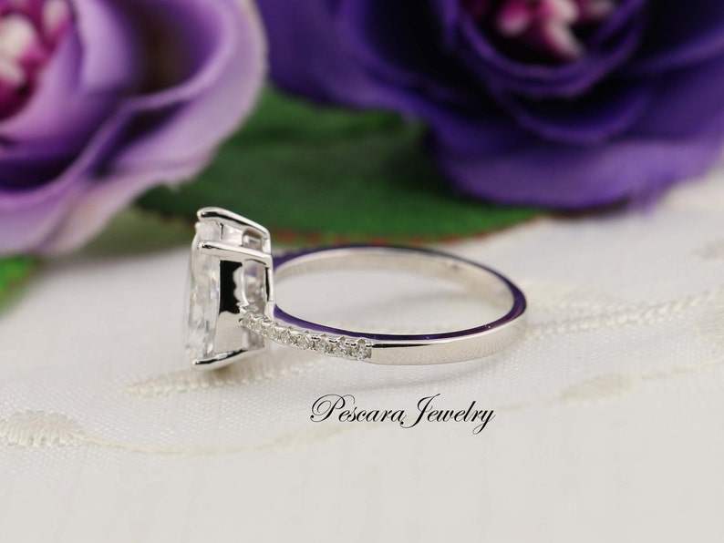 May include: A silver ring with a pear-shaped clear stone and a band of smaller clear stones. The ring is on a white surface with a purple flower in the background.