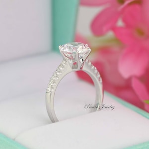 May include: A silver engagement ring with a large, clear, round-cut center stone and a band of smaller, clear stones. The ring is in a light green box with pink flowers in the background.