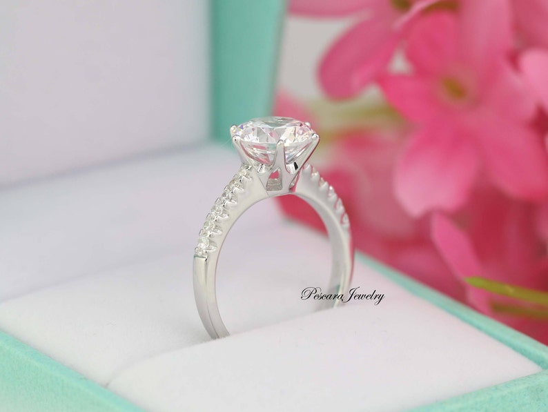 May include: A silver engagement ring with a large, clear, round-cut center stone and a band of smaller, clear stones. The ring is in a light green box with pink flowers in the background.