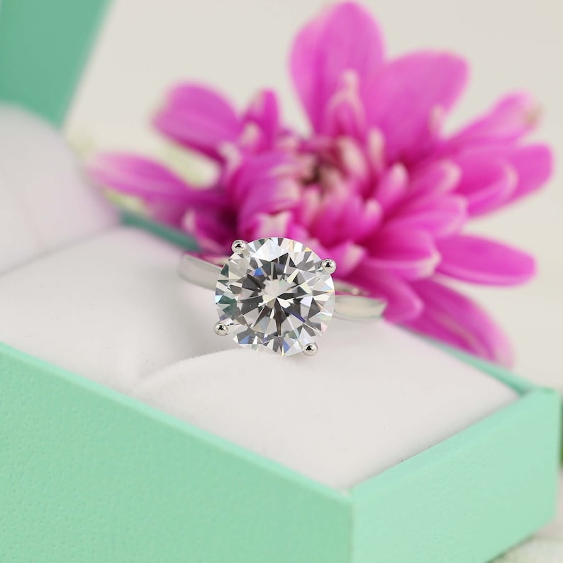May include: A sparkling diamond engagement ring set in a silver band, displayed in a light green ring box with a pink flower.