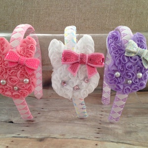 Puede incluir: Tres diademas de conejito de color pastel con rosetas de tela blanca, rosa y morada. Cada diadema tiene un lazo con lentejuelas y un centro blanco perlado.