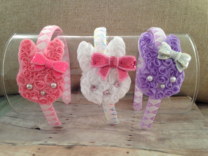 Puede incluir: Tres diademas de conejito de color pastel con rosetas de tela blanca, rosa y morada. Cada diadema tiene un lazo con lentejuelas y un centro blanco perlado.