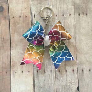 May include: A rainbow mermaid-scale patterned bow with a silver key chain. The bow is tied with a white ribbon.