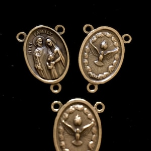 May include: Three silver-toned metal charms with religious imagery. The first charm features the Holy Family, the second charm features a dove, and the third charm features a dove with a flame above it.