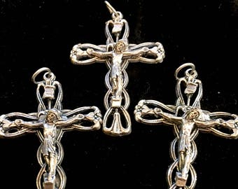 Op de afbeelding: Drie zilverkleurige kruisvormige hangers. Elk kruis heeft een gedetailleerde afbeelding van Jezus Christus en sierlijke bloemen- en lusontwerpen. De hangers worden gepresenteerd tegen een zwarte achtergrond, waardoor hun ingewikkelde details worden benadrukt.