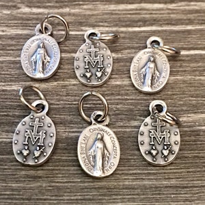 May include: Six silver charms with a religious theme. The charms feature the Virgin Mary and the words "Regina Sine Labe Originalli Concepta" which translates to "Queen Without Stain Originally Conceived".