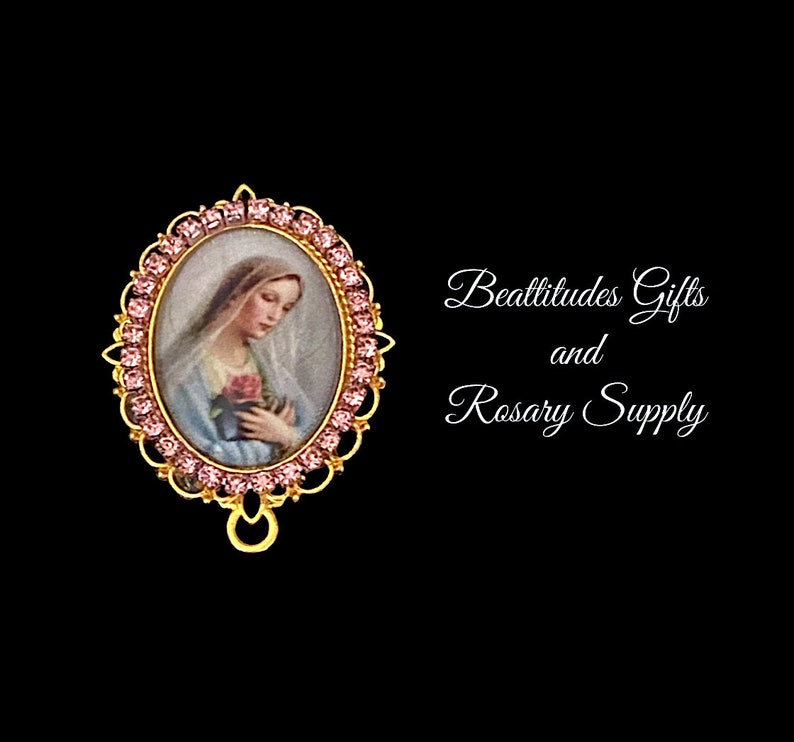 Puede incluir: Un colgante ovalado en tono dorado con un borde de strass rosa. El colgante presenta una imagen de la Virgen Mar&iacute;a sosteniendo una rosa. El texto "Beatitudes Gifts and Rosary Supply" es visible en el fondo.