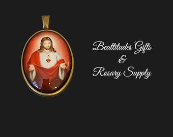 May include: A gold oval pendant with a picture of Jesus Christ with his heart exposed. The pendant is on a black background with the text "Beattitudes Gifts & Rosary Supply".