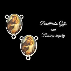 May include: Two silver-toned oval-shaped charms with a picture of Saint Anthony of Padua holding the baby Jesus. The charms have three loops for attaching to a rosary or necklace. The text "Beatitudes Gifts and Rosary supply" is visible in the background.