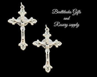 May include: Two silver-toned crucifix pendants with ornate detailing, featuring the figure of Jesus Christ. The pendants are suspended against a black background. The text "Beattitudes Gifts and Rosary supply" is written in cursive.
