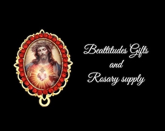 May include: An oval pendant with a gold frame and red gemstones. The pendant features an image of Jesus with a heart and a crown of thorns. The text "Beattitudes Gifts and Rosary supply" is on the right side of the image.