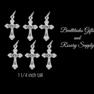 May include: Six silver-colored crucifix pendants, each featuring a detailed depiction of Jesus on the cross. The pendants are 1 1/4 inches tall and have a loop at the top for attaching to a chain. The text "Beattitudes Gifts and Rosary Supply" is also visible.