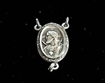 May include: Silver-toned metal religious charm featuring a detailed image of Jesus Christ with a heart on his chest. The charm is oval-shaped and has a decorative border.