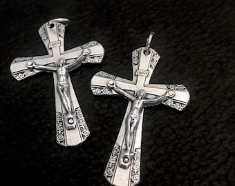 May include: Two silver-colored crucifix pendants. Each cross features a detailed depiction of Jesus Christ and the inscription "INRI" at the top. The crosses have decorative floral patterns and are displayed on a dark background.