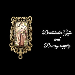 May include: A gold-toned metal frame with an ornate design surrounds a rectangular image of a woman in a white habit holding a bouquet of roses. The text "Beatitudes Gifts and Rosary supply" is written in white cursive script on a black background.