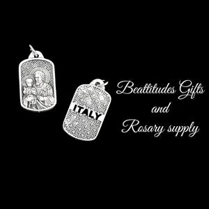 Op de afbeelding: Twee zilveren metalen hangers, een met een religieus beeld van een man en een kind, en de andere met het woord "ITALI&Euml;" in een decoratief lettertype. De tekst "Beatitudes Gifts and Rosary supply" is geschreven in wit cursief schrift op een zwarte achtergrond.