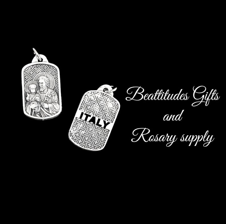 Op de afbeelding: Twee zilveren metalen hangers, een met een religieus beeld van een man en een kind, en de andere met het woord "ITALI&Euml;" in een decoratief lettertype. De tekst "Beatitudes Gifts and Rosary supply" is geschreven in wit cursief schrift op een zwarte achtergrond.