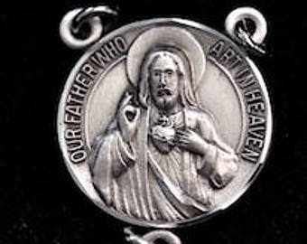 May include: Silver-toned metal charm featuring a depiction of Jesus Christ with a heart on his chest. The charm has the words "Our Father Who Art In Heaven" inscribed around the edge.