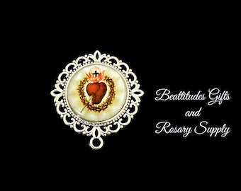 May include: Silver-toned pendant with a white background and a red heart with a crown of thorns and a cross. The pendant is surrounded by a decorative silver frame. The text "Beatitudes Gifts and Rosary Supply" is visible in the background.