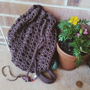 May include: A brown crocheted bag with a drawstring closure and colourful beads. The bag is lying on a concrete surface next to a potted plant with yellow flowers.